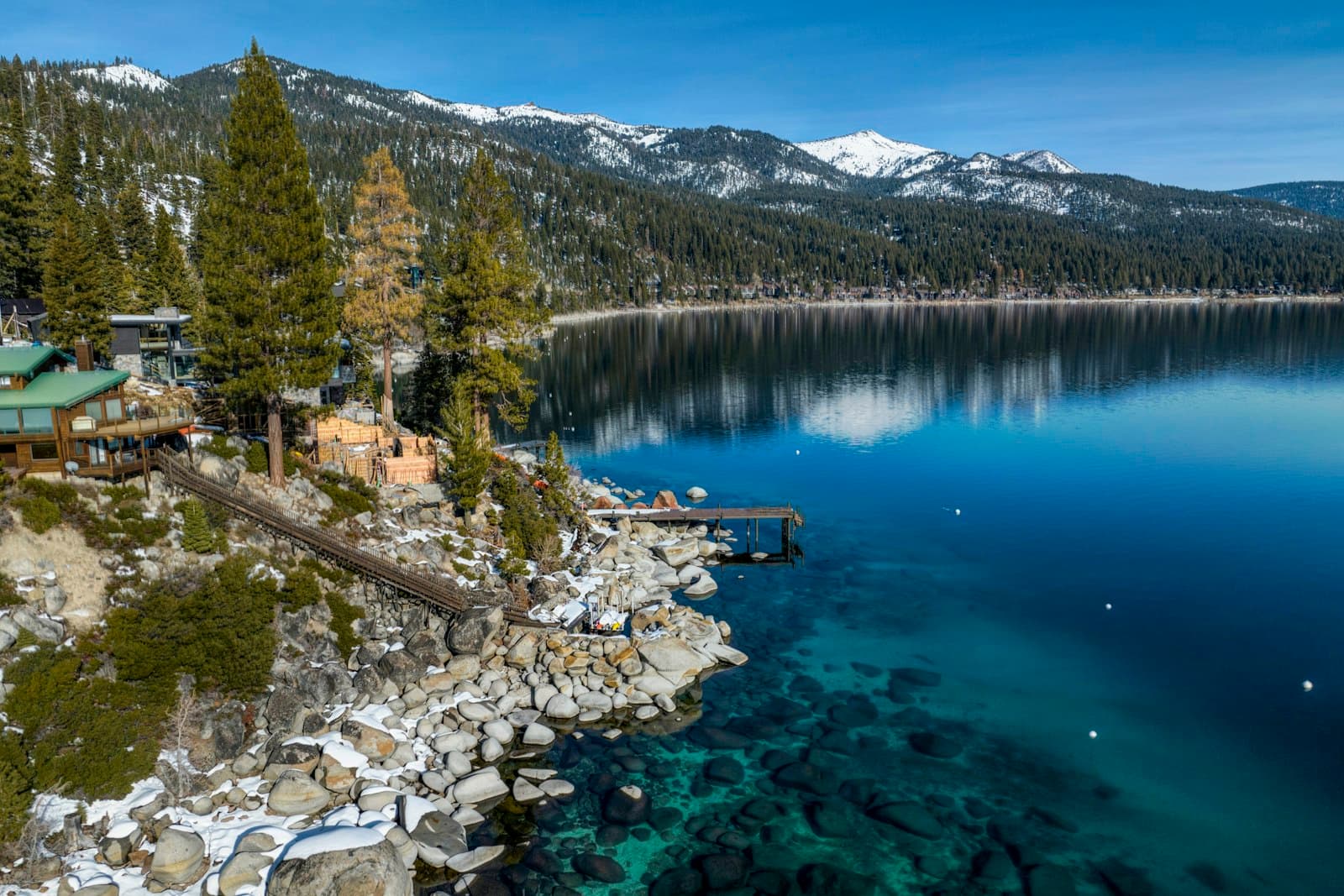 Lakefront homes and a private pier among pines on the clear blue water of Lake Tahoe, with snow-capped mountains behind.