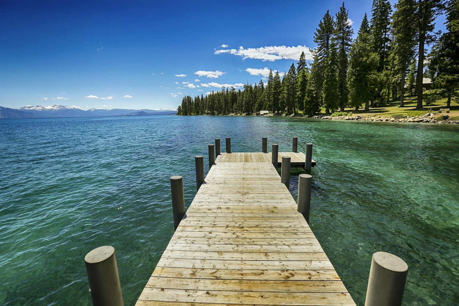 A wooden pier reaching into the clear green water of Lake Tahoe on a sunny day, with pines along the shore.