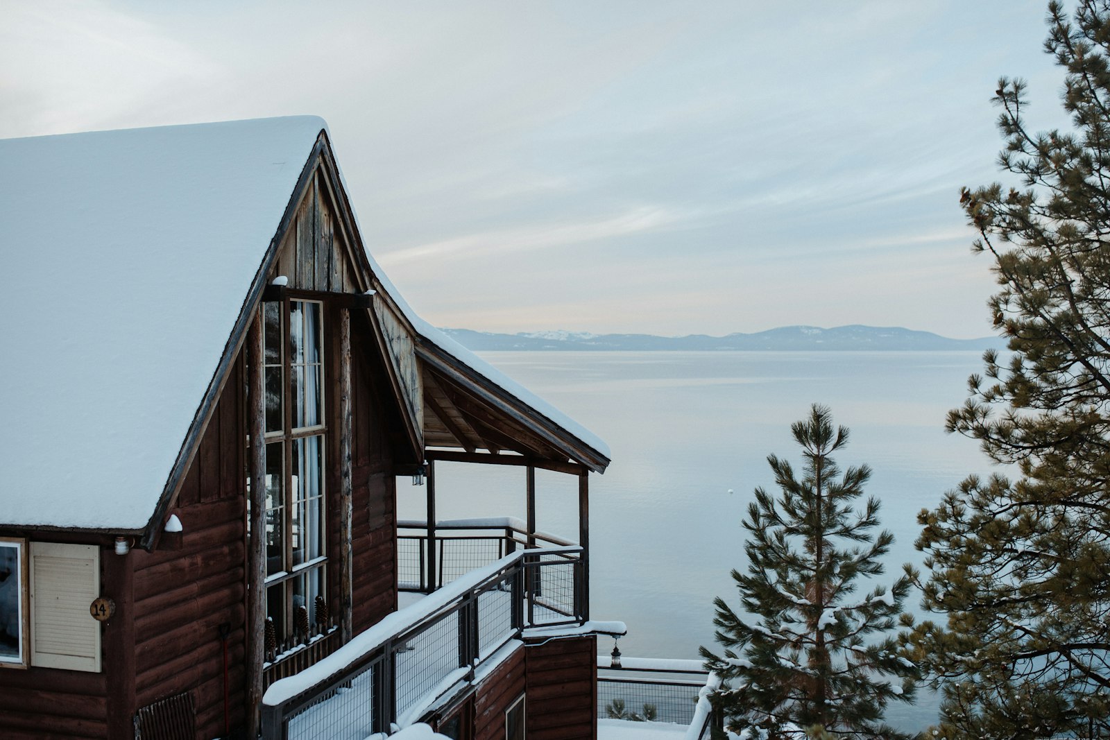 A wood-sided cabin with a snow-covered roof and a deck looking out over Lake Tahoe through the pines.
