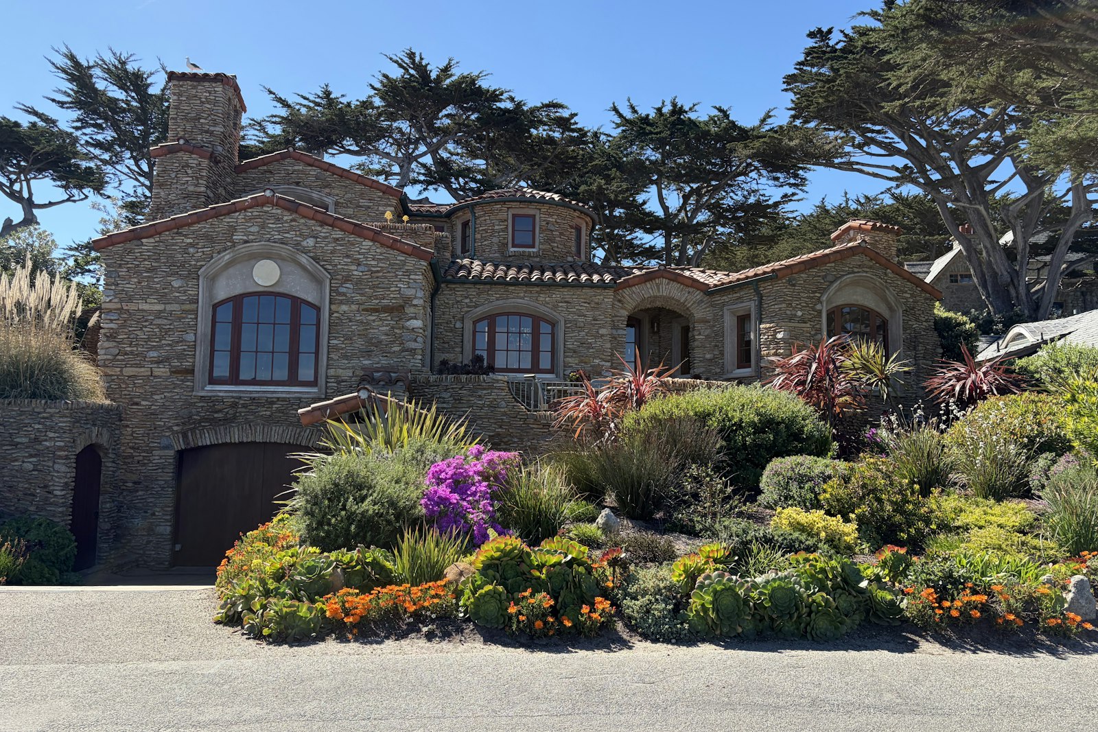 A stone house with arched windows behind a flowering front garden and tall cypress trees on the Monterey Peninsula.